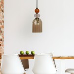 Lampa wisząca Tropea z kloszem w kolorze bursztynowym i czarnym, zawieszona na czarnym przewodzie, z dekoracyjnymi, kolorowymi kulami. Widoczna nad drewnianym stołem z białymi krzesłami i zielonymi jabłkami. Tło jasne, minimalistyczne.