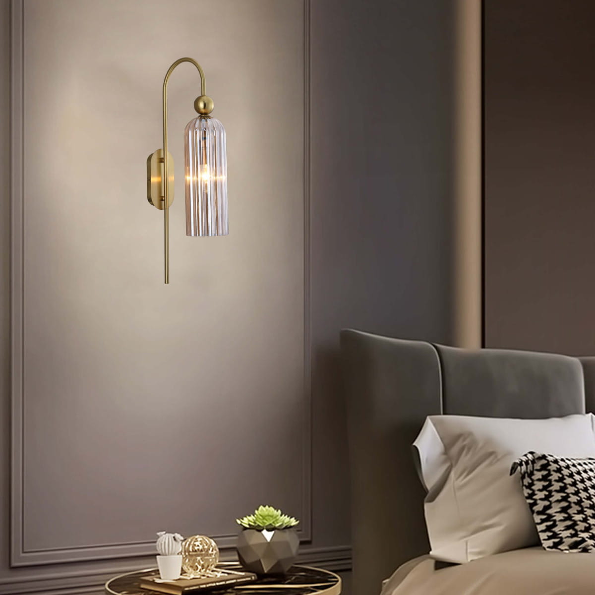 Kinkiet Piega z patynowym wykończeniem, z bursztynowym kloszem z przezroczystego szkła, umieszczony na ścianie. Oświetlenie o eleganckim designie, z widocznym źródłem światła, w aranżacji sypialni obok łóżka i dekoracyjnych akcesoriów.