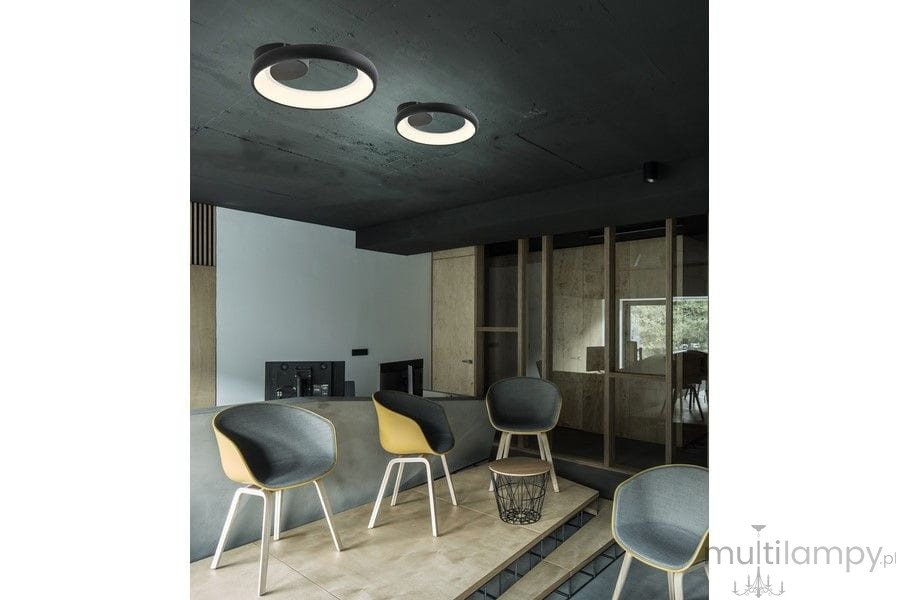 Czarna lampa sufitowa Thea z jedną żarówką LED 20W, o okrągłym kształcie, zamontowana na ciemnym suficie. W tle nowoczesne wnętrze z minimalistycznymi krzesłami i drewnianymi akcentami.