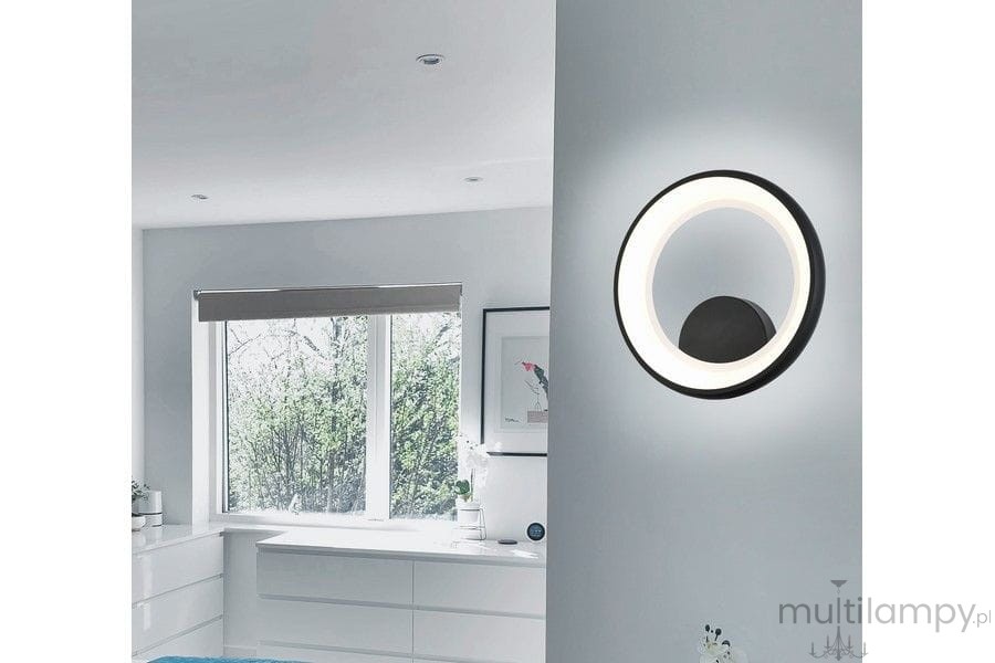Kinkiet Thea w kolorze czarnym z 1xLED 20W, zamontowany na ścianie. Okrągły design z białą obręczą emitującą światło, w tle jasne wnętrze z oknem i meblami.
