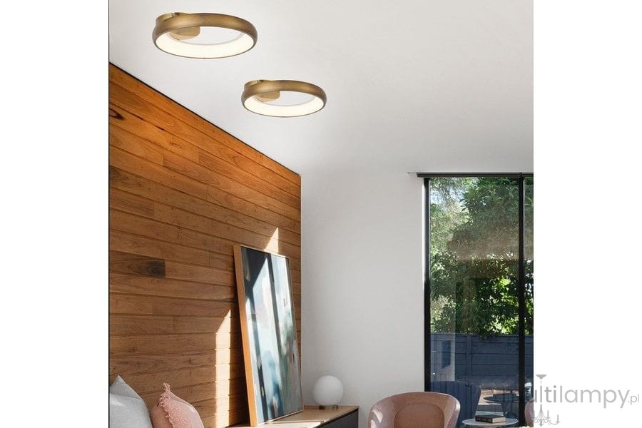 Lampa sufitowa Thea w kolorze złotym, pojedyncza, z technologią LED o mocy 20W. Zamontowana na białym suficie, z delikatnym okrągłym kloszem. Widoczna w nowoczesnym wnętrzu z drewnianą ścianą i meblami w stonowanych kolorach.