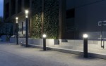 Lampa stojąca ogrodowa Barel H98, kolor ciemny popiel, wykonana z metalu. Posiada źródło światła LED 23W. Ustawiona pionowo na schodach, emitując ciepłe światło, w towarzystwie roślinności oraz nowoczesnej architektury w tle.