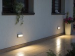 Szara lampa elewacyjna Mur PC LED, zamontowana na ścianie, emitująca ciepłe światło. W tle widoczna donica z kwiatami oraz roślinność na parapecie. Oświetlenie podłogi wzdłuż ściany, na jasnych płytkach.