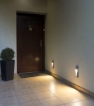 Szara lampa elewacyjna LED Mur PC 2502B zamontowana na ścianie, emitująca ciepłe światło. W tle widoczna drewniana drzwi oraz donica z rośliną. Lampa w pozycji pionowej, na tle jasnej ściany i płytek podłogowych.