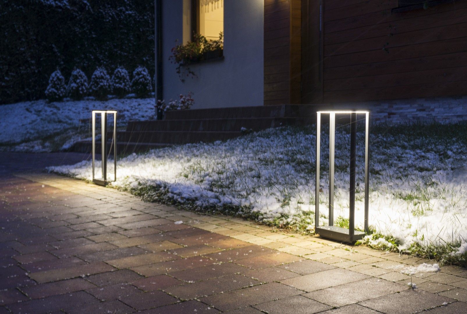Lampa stojąca Arma H50 w ciemnym popiele, z jedną żarówką LED o mocy 9W. Ujęcie z boku, pokazujące lampę w oświetlonym otoczeniu, na tle trawnika pokrytego śniegiem oraz kostki brukowej.