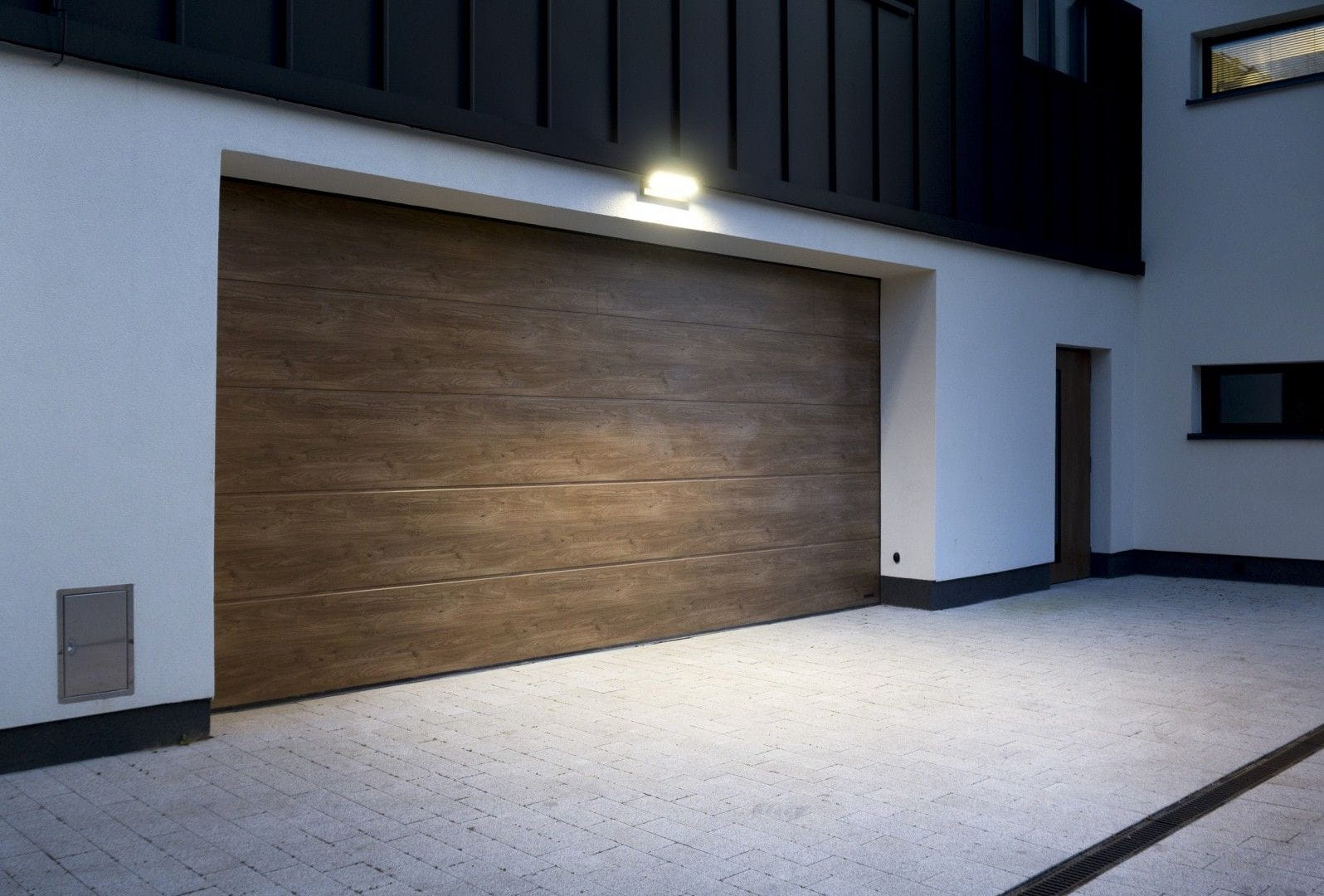 Kinkiet elewacyjny kierunkowy LED 27W 4000K w kolorze ciemnego popielu, zamontowany na ścianie, oświetlający wjazd. Prosty kształt, umieszczony nad bramą, z czujnikiem ruchu. Tło w jasnych odcieniach z płytkami i drewnianą bramą.
