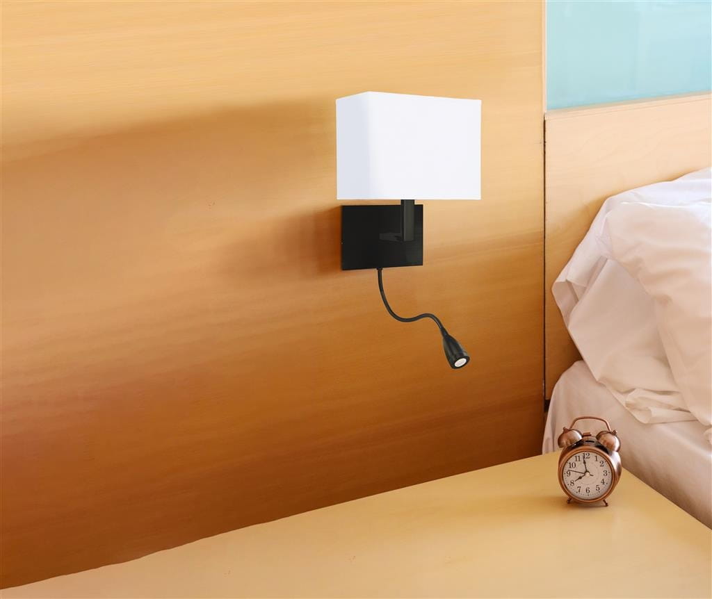 Kinkiet hotelowy w kolorze czarnym z białym prostokątnym kloszem oraz dodatkowym elastycznym ramieniem z małą lampką. Zamontowany na ścianie obok łóżka, w tle widoczny stół nocny z zegarem.