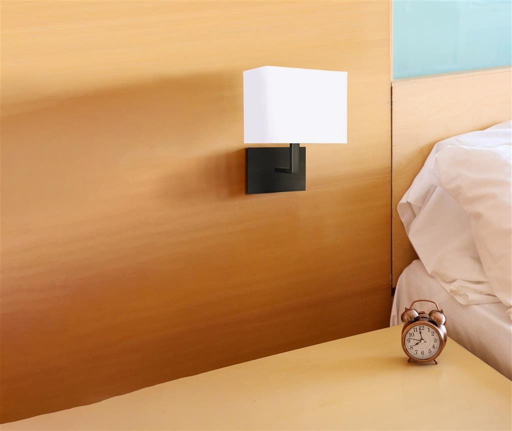Kinkiet hotelowy 1-punktowy w kolorze czarnym z prostokątnym białym kloszem, zamontowany na ścianie obok łóżka. Na stoliku nocnym znajduje się budzik w stylu retro, a w tle widoczna jest jasna ściana z drewnianymi elementami.