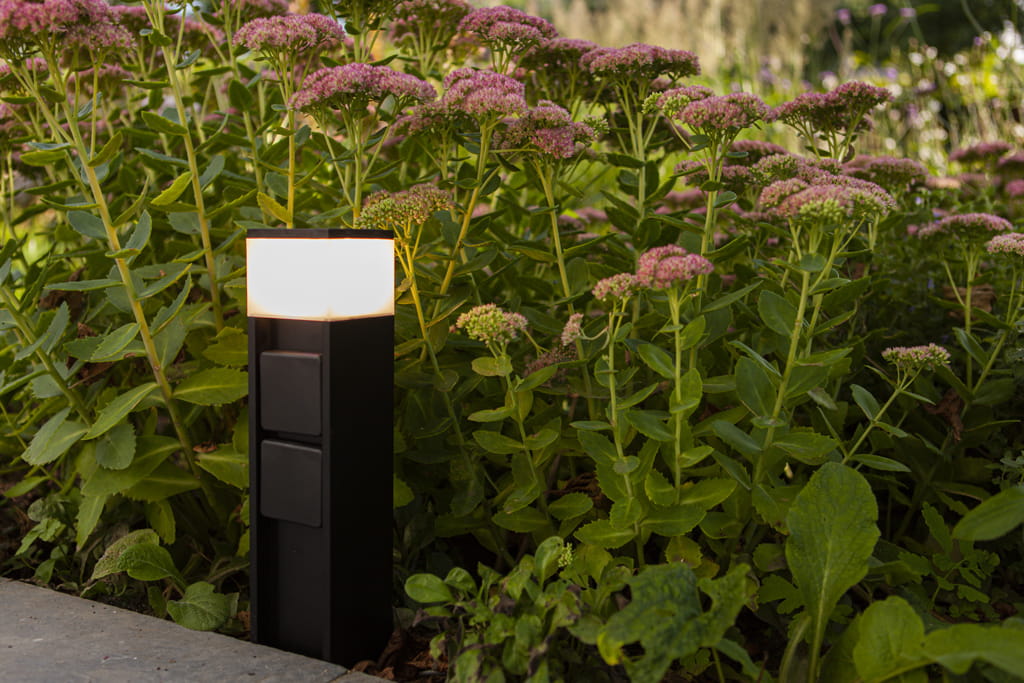 Słupek ogrodowy w kolorze czarnym z wbudowanymi dwoma gniazdami typu F i lampą LED 11W. Ujęcie z boku, umieszczony w zielonym otoczeniu, wśród kwiatów. Górna część z mlecznym kloszem, dolna z widocznymi gniazdami.
