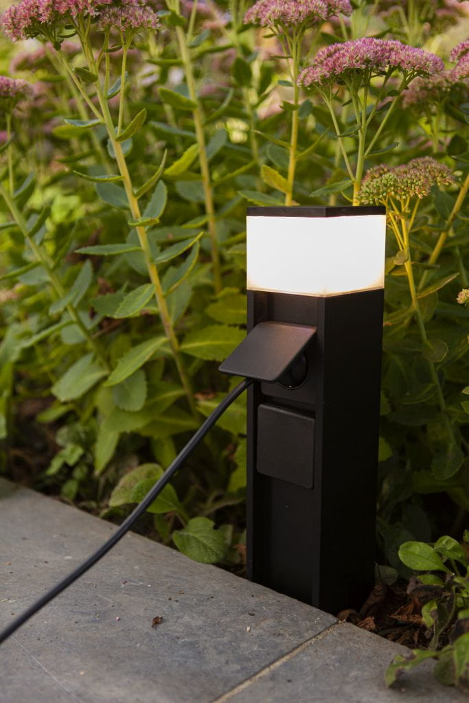 Słupek ogrodowy Mains w kolorze czarnym z 2 wbudowanymi gniazdami typu F oraz lampą LED 11W. Ujęcie z boku, z widocznym złączem i kablem, otoczony zielenią roślin. Górna część z mlecznego tworzywa, emitującego światło.