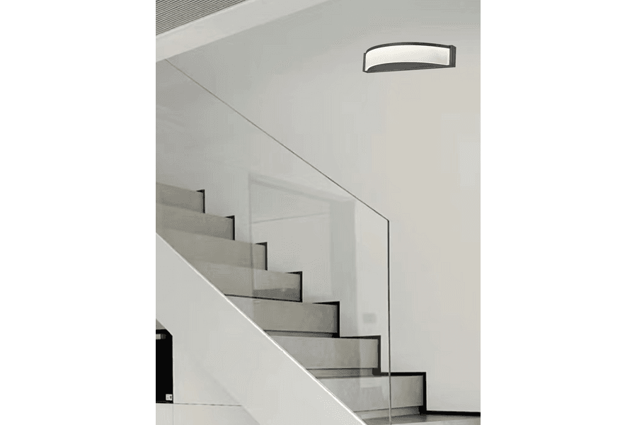 Kinkiet Cornelia 1xLED/10W w kolorze czarnym, zamontowany na ścianie obok schodów. Posiada półokrągły kształt z białym kloszem, emitującym światło. Tło jasne, minimalistyczne, podkreślające nowoczesny design lampy.