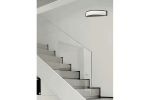 Kinkiet Cornelia 1xLED/10W w kolorze czarnym, zamontowany na ścianie obok schodów. Posiada półokrągły kształt z białym kloszem, emitującym światło. Tło jasne, minimalistyczne, podkreślające nowoczesny design lampy.