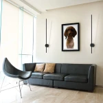 Kinkiet Chloe 85 w kolorze czarnym, z jedną diodą LED o mocy 11W i temperaturze barwowej 3000K. Zamontowany na ścianie, minimalistyczny design z prostokątnym źródłem światła oraz okrągłym mocowaniem. W tle widoczna kanapa i obraz.