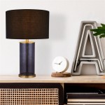 Lampa stołowa Liberty z mosiężnym wykończeniem i czarnym abażurem, o cylindrycznym kształcie z przezroczystym korpusem. Ujęcie frontalne, lampa stoi na drewnianym stoliku obok dekoracyjnego zegara i litery „A” w tle.