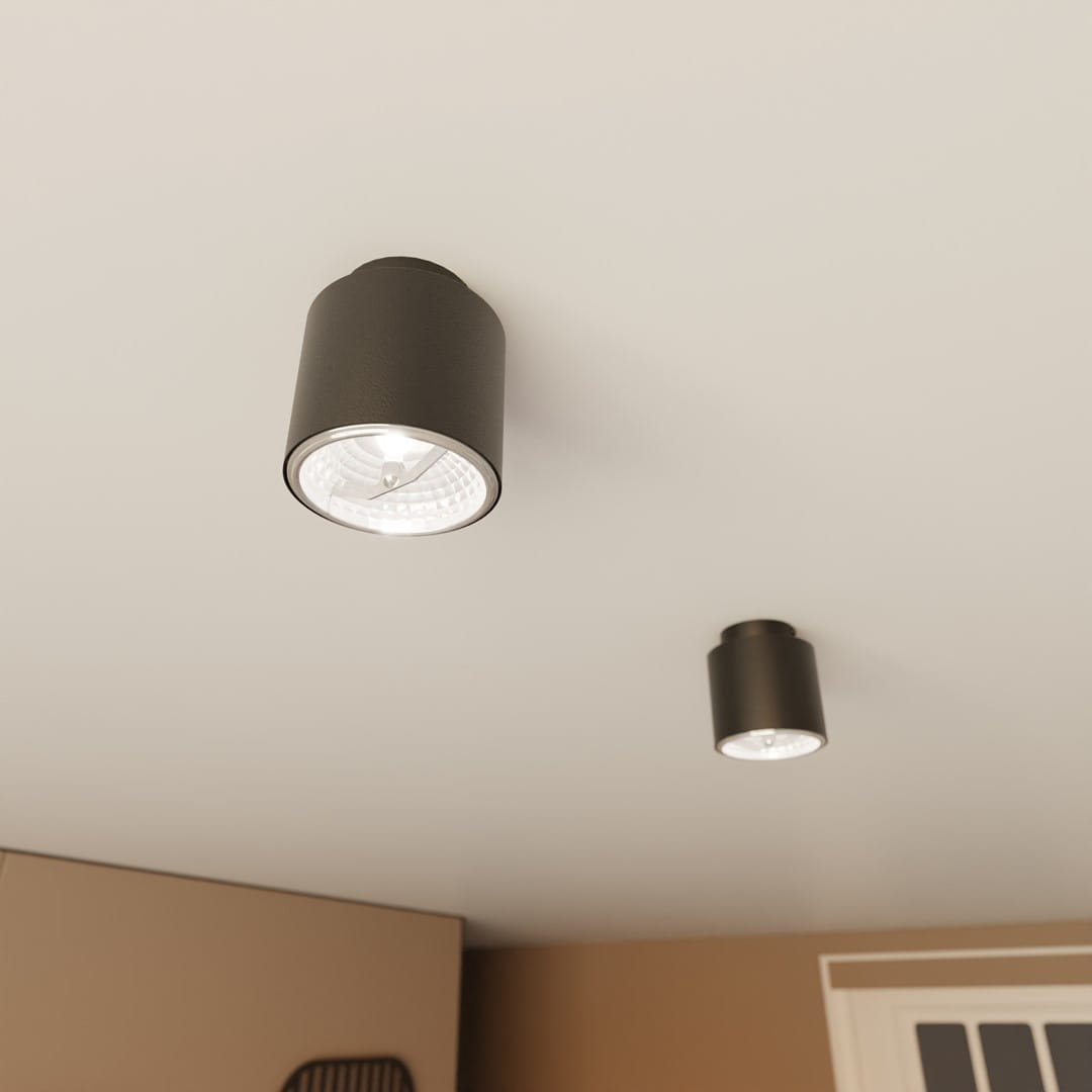Czarna nano lampa sufitowa z jedną oprawą GU10/AR 111, o cylindrycznym kształcie. Ujęcie z dołu, ukazujące lampę zamontowaną na białym suficie. W tle widoczna druga lampa o podobnym designie. Jasne światło emitowane z wnętrza lampy.