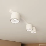 Biała lampa sufitowa z trzema źródłami światła GU10, zamontowana na prostokątnej podstawie. Klosze w formie cylindrów, z metalicznymi elementami, skierowane w różne strony. Minimalistyczny design, idealna do nowoczesnych wnętrz.
