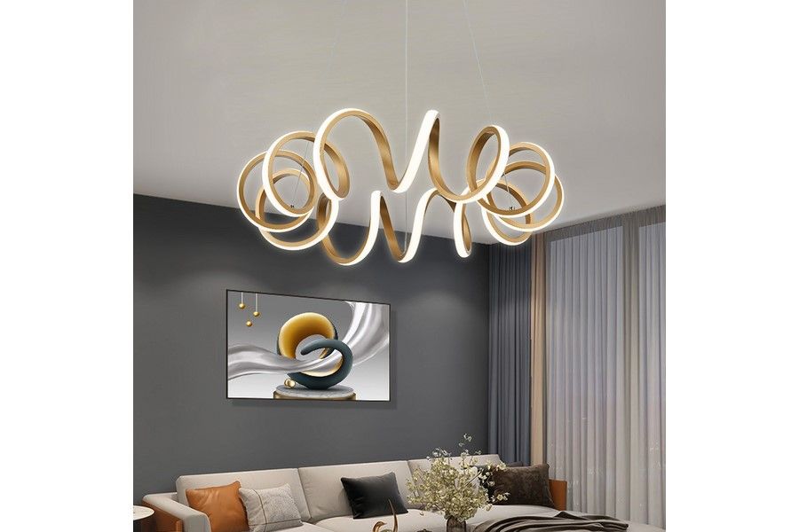 Lampa wisząca Loop w kolorze złotym z pilotem, posiadająca unikalny, falisty kształt. Wykonana z materiałów LED, emitująca ciepłe światło. Zawieszona w nowoczesnym wnętrzu, na tle szarej ściany i zasłon.