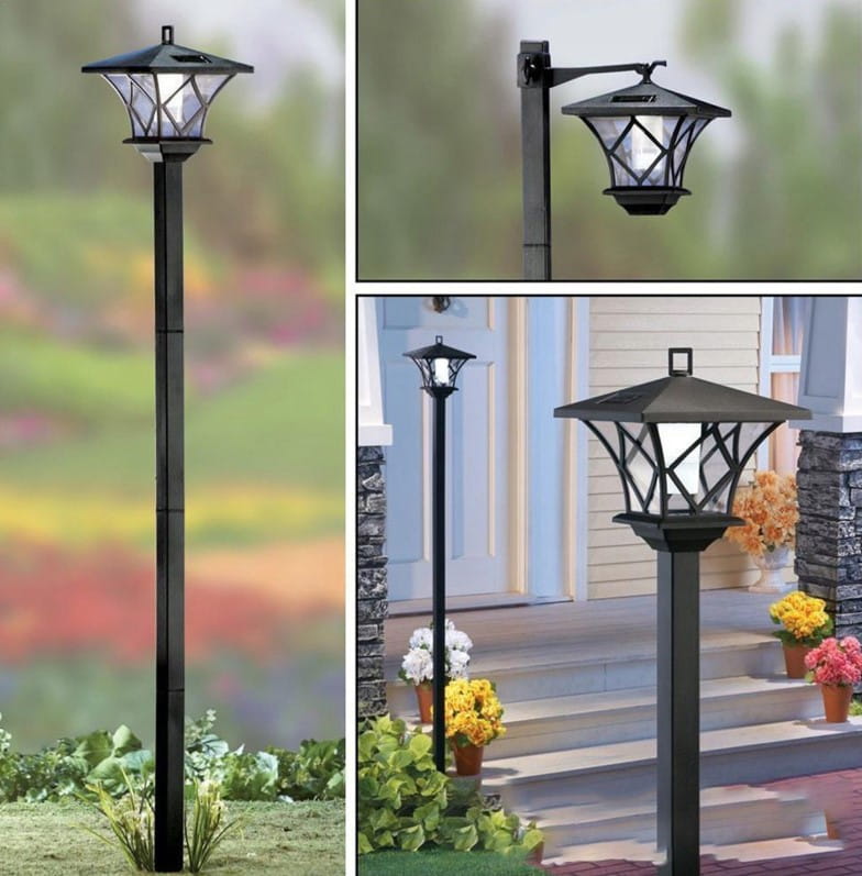 Lampa solarna ogrodowa z czujnikiem zmierzchu, czarna. Wysoka, na prostej podstawie, z dekoracyjnym kloszem w kształcie heksagonu. W tle widoczne kwiaty i zielona trawa, lampa ustawiona w ogrodzie.