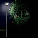 Lampa solarna ogrodowa z czujnikiem zmierzchu LED, czarna, o wysokim słupku. Wzór klosza przypominający latarnię, z dekoracyjnymi elementami. W tle widoczne rośliny, co podkreśla jej zastosowanie w ogrodzie.