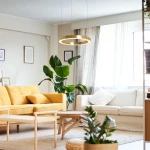 Lampa wisząca Aurum D60 w kolorze złotym, z jednym źródłem LED o mocy 27W. Umieszczona w nowoczesnym wnętrzu nad drewnianym stołem, otoczona roślinami i meblami w jasnych odcieniach. Kąt ujęcia z boku, ukazujący elegancki design lampy.