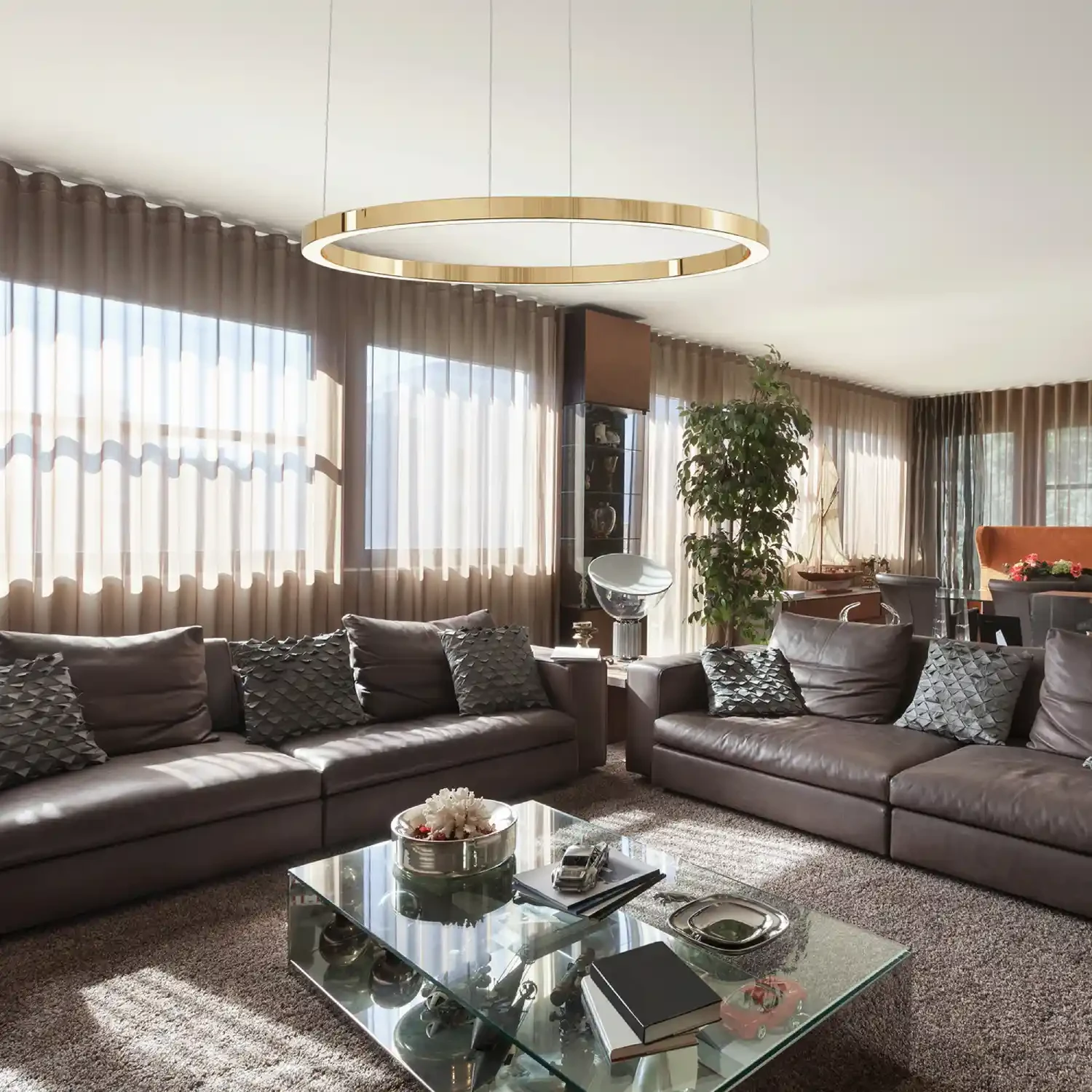 Lampa wisząca Aurum D120 w kolorze złotym, z pojedynczym źródłem światła LED o mocy 55W. Zawieszona na cienkich linkach, z okrągłym kloszem, w nowoczesnym wnętrzu z brązowymi sofami i szklanym stolikiem.