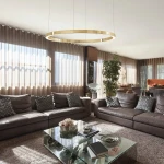 Lampa wisząca Aurum D120 w kolorze złotym, z pojedynczym źródłem światła LED o mocy 55W. Zawieszona na cienkich linkach, z okrągłym kloszem, w nowoczesnym wnętrzu z brązowymi sofami i szklanym stolikiem.