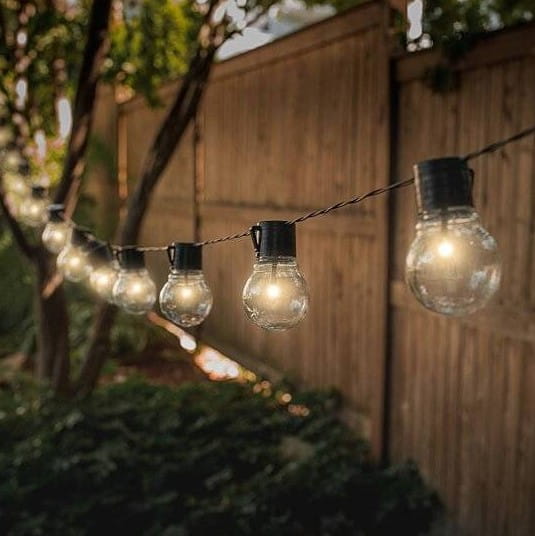 Girlanda ogrodowa solarna z 30 kulkami LED, długość 7,7 m, z czarnym przewodem. Kule LED w kształcie żarówek zawieszone w równych odstępach, otoczone zielenią, w tle drewniana ogrodzenie.