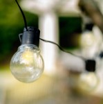 Girlanda ogrodowa solarna z 30 kulkami LED, długość 7,7 m, czarna. Widoczne szklane kulki w formie żarówek z czarnymi oprawkami, zawieszone na cienkim przewodzie. Tło rozmyte, z elementami zieleni.