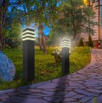 Lampa stojąca Malibu H50, kwadratowa, czarna, z jedną oprawą E27. Widoczna w ogrodzie, oświetlająca ścieżkę, z przezroczystymi elementami u góry. Ustawiona na tle trawnika i drzew, w otoczeniu kamieni i roślin.