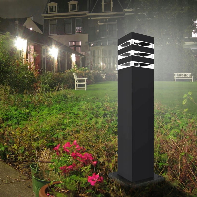 Lampa stojąca Malibu H50 w kształcie kwadratu, czarna, z jedną oprawą E27. Ujęcie z boku, prezentująca proste linie i szczeliny w górnej części, w ogrodzie otoczona zielenią i kwiatami. Idealna do oświetlenia przestrzeni zewnętrznych.