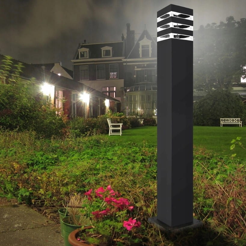 Czarna lampa stojąca słupek Malibu H80 o kwadratowym kształcie, z jedną oprawą E27. Ujęcie z boku, w ogrodzie, z widocznymi elementami otoczenia, takimi jak trawa i kwiaty. Lampa o nowoczesnym designie z dekoracyjnymi szczelinami na górze.