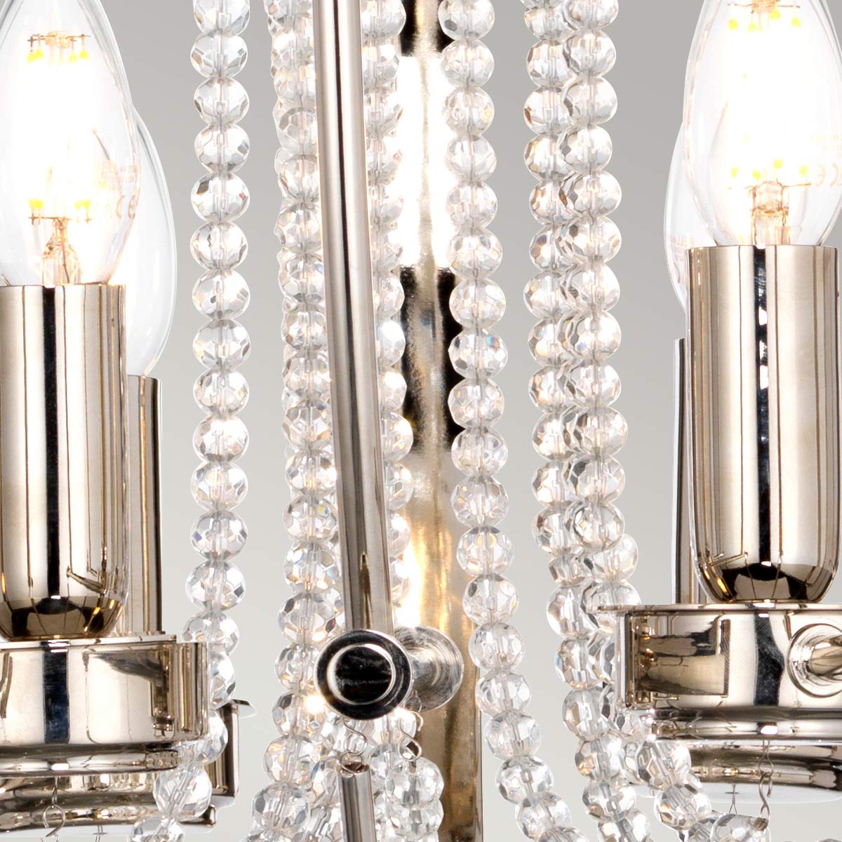 Lampa wisząca Cassie z polerowanego niklu, z sześcioma gniazdami E14 i jednym E27. W centralnej części metalowa konstrukcja, otoczona kryształowymi koralikami, które odbijają światło. Kąt ujęcia zbliżeniowy, ukazujący detale wykończenia.