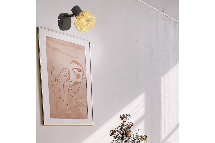 Kinkiet Loretto w kolorze czarnym i złotym, z pojedynczym źródłem światła E14, zamontowany na ścianie obok abstrakcyjnego obrazu. Okrągła, siatkowa osłona lampy emitująca ciepłe światło, na tle jasnej ściany i dekoracyjnych elementów.