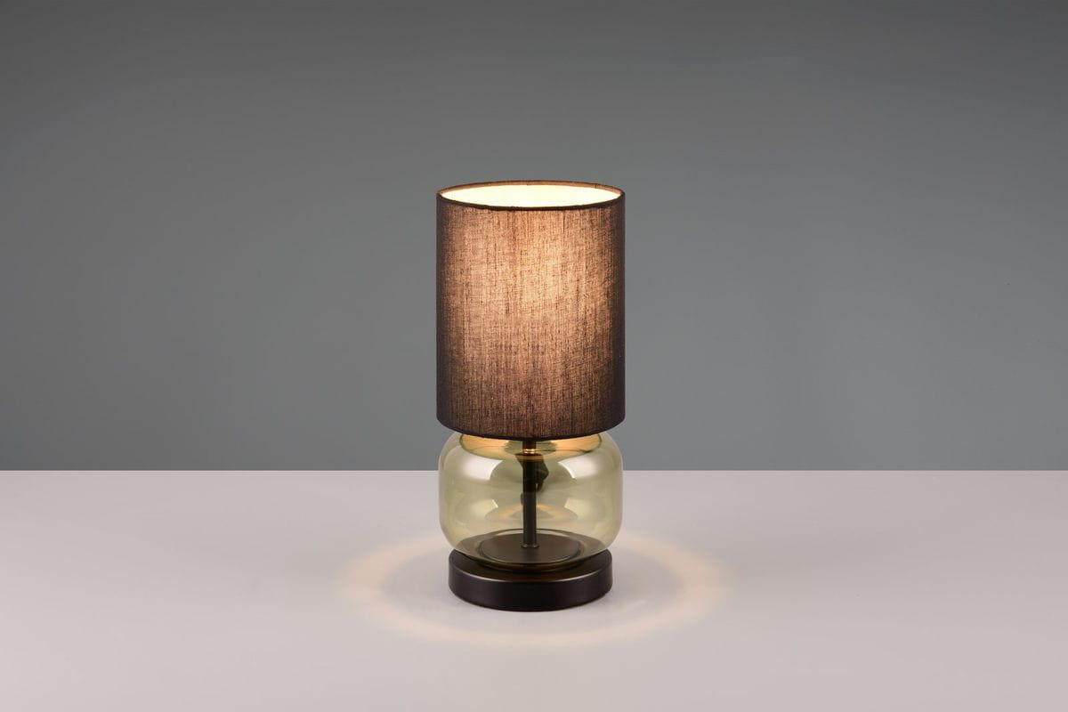 Lampa stołowa Elio, z kloszem w kolorze czarnym i podstawą z zielonego szkła, stojąca na okrągłej, ciemnej podstawie. Klosz wykonany z materiału tekstylnego, emitujący ciepłe światło. Ujęcie frontalne, na jednolitym tle.