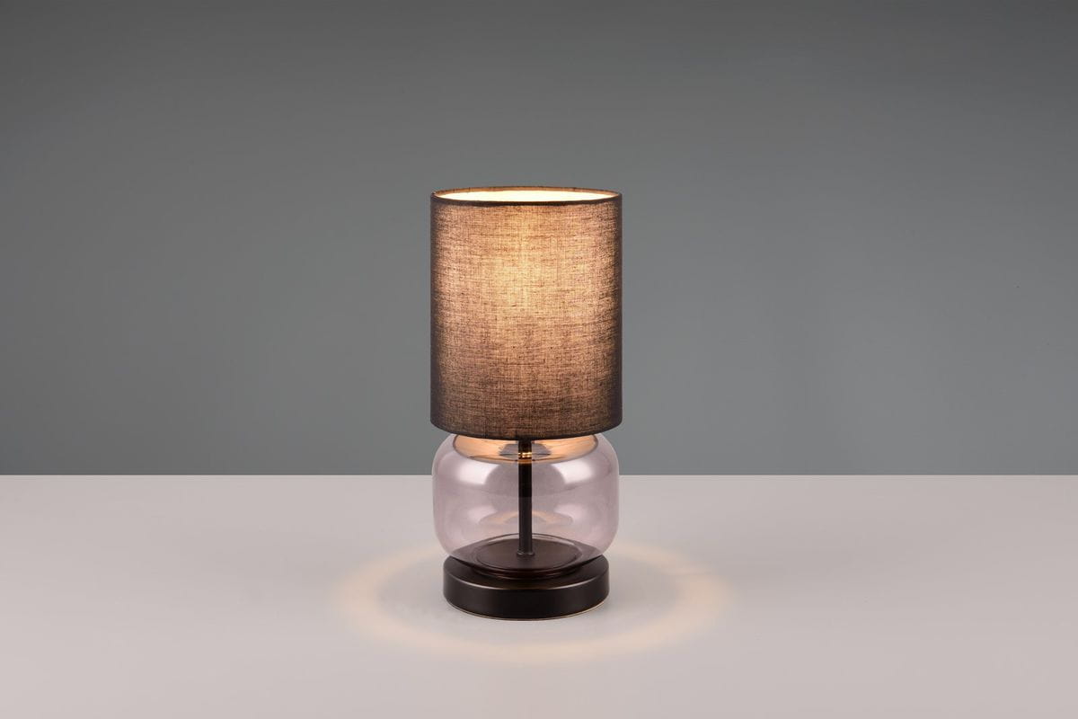 Lampa stołowa Elio 1xE27 w przydymionej szklanej podstawie i czarnym abażurze. Ujęcie frontalne, stojąca na białym blacie. Abażur o okrągłym kształcie, materiał tekstylny, emitujący ciepłe światło. Podstawa okrągła, ciemna, z metalowym elementem.