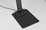 Lampka biurkowa Travis w kolorze czarnym z portem USB-C. Posiada prostokątną podstawę z dotykowym panelem sterowania, zawierającym przycisk zasilania oraz regulację jasności. Kąt ujęcia ukazuje dolną część lampy i jej detale.
