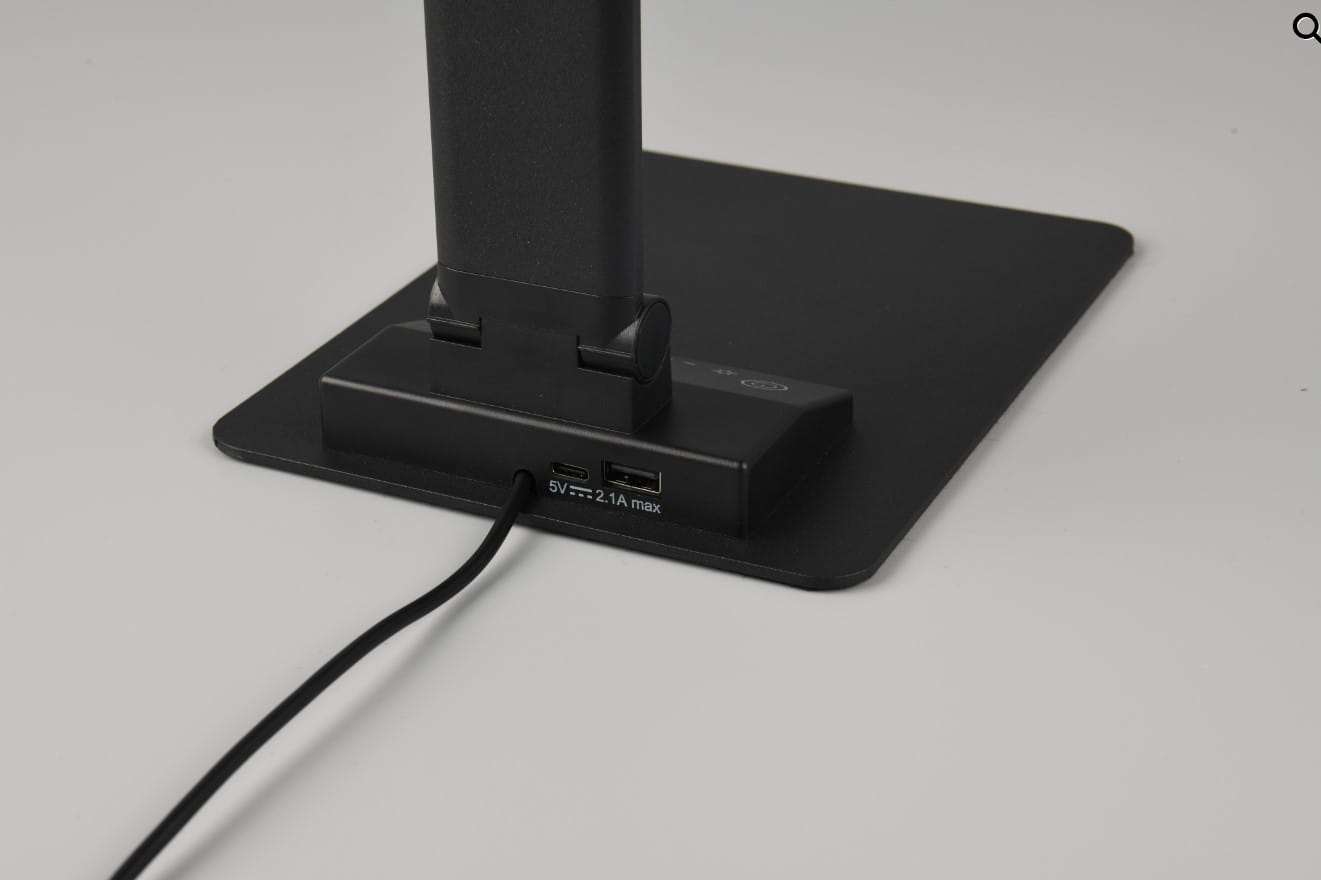 Czarna lampka biurkowa Travis z portem USB-C, wykonana z materiału matowego, z prostokątną podstawą. Widoczne dwa porty USB oraz kabel zasilający. Ujęcie z boku, prezentujące detale w dolnej części lampki.