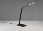 Lampka biurkowa Travis w kolorze czarnym z portem USB-C, wykonana z metalu, z prostokątnym kloszem LED o mocy 6,5W. Ujęcie z boku, stojąca na płaskiej podstawie, z widocznym kablem zasilającym. Tło jednolite, szare.