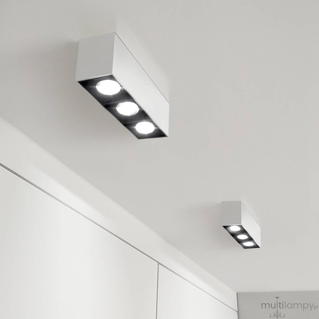 Lampa sufitowa LED w kolorze białym, z trzema okrągłymi źródłami światła, zamontowana na suficie. Prosty, prostokątny design. Widoczne również mniejsza lampa w tle, utrzymana w tym samym stylu.