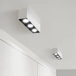 Lampa sufitowa LED w kolorze białym, z trzema okrągłymi źródłami światła, zamontowana na suficie. Prosty, prostokątny design. Widoczne również mniejsza lampa w tle, utrzymana w tym samym stylu.