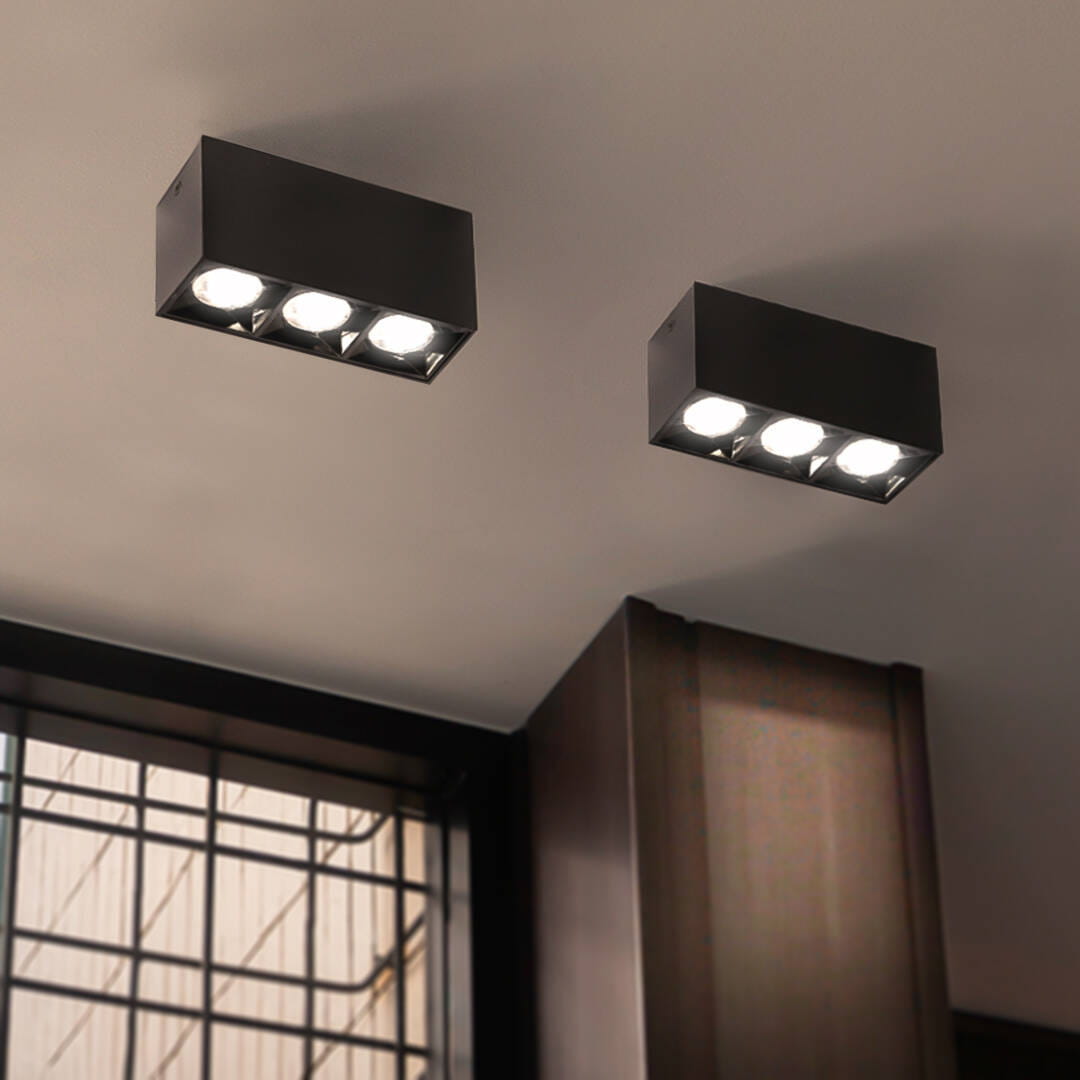 Lampa sufitowa LED o mocy 9W i temperaturze barwowej 4000K, czarna, zamontowana na suficie. Posiada dwa prostokątne korpusy z trzema okrągłymi źródłami światła w każdym. Tło stanowi gładka powierzchnia sufitu.