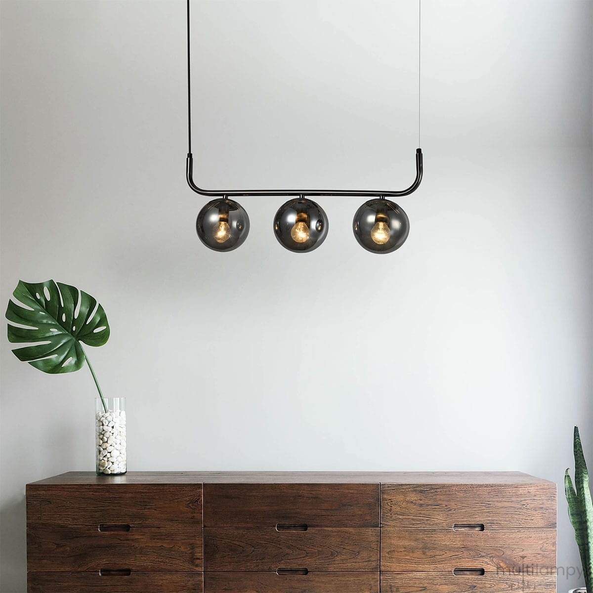 Lampa wisząca Alisa 3XE14 w kolorze grafitowym z trzema dymionymi kloszami, zawieszona na prostej metalowej konstrukcji. W tle drewniana komoda i roślina w doniczce, na jasnym tle.