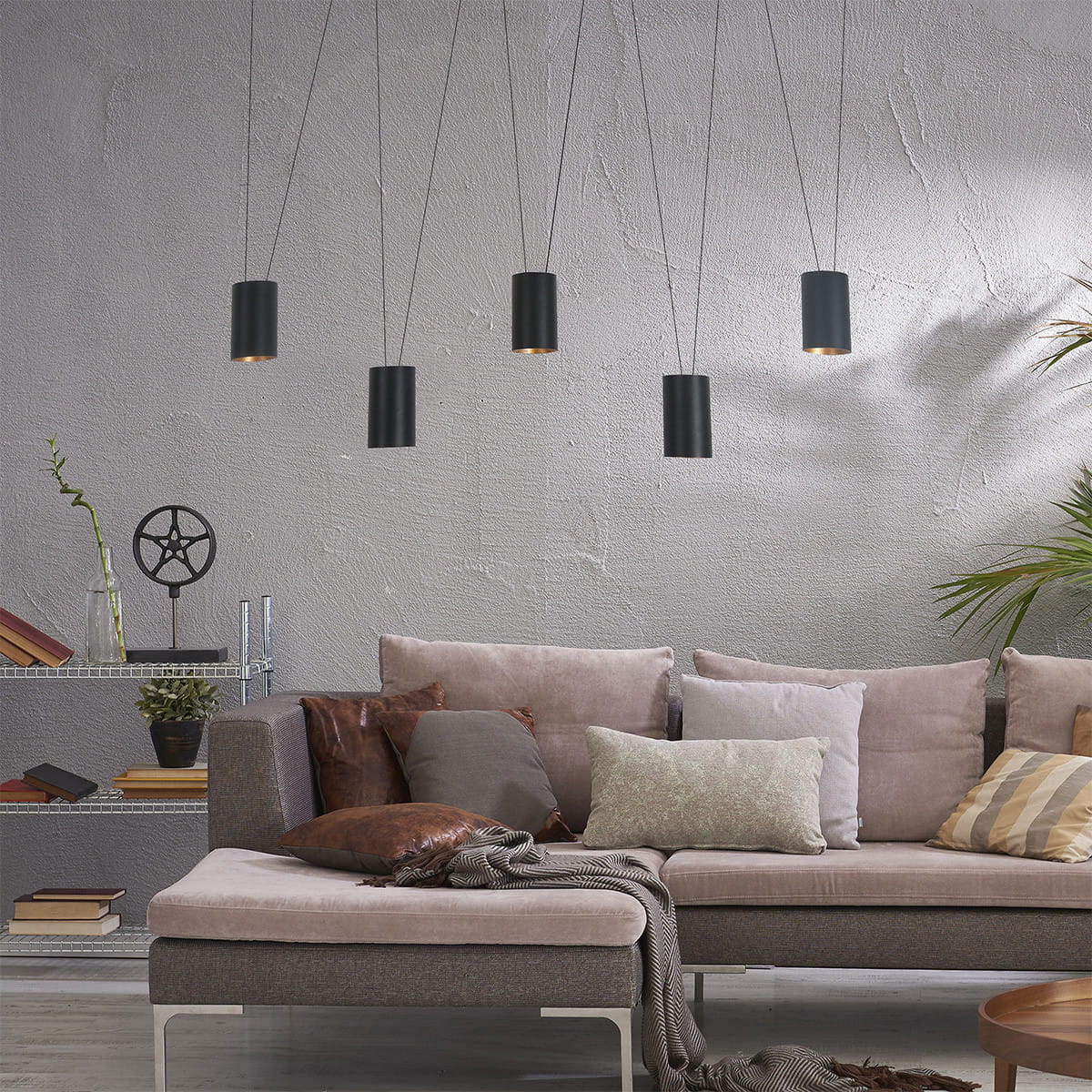 Lampa wisząca Toledo z pięcioma cylindrycznymi kloszami w kolorze czarnym, zawieszona na cienkich linkach. W tle kanapa z poduszkami w różnych kolorach, na szarej ścianie oraz regał z książkami i roślinnością.