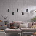 Lampa wisząca Toledo z pięcioma cylindrycznymi kloszami w kolorze czarnym, zawieszona na cienkich linkach. W tle kanapa z poduszkami w różnych kolorach, na szarej ścianie oraz regał z książkami i roślinnością.
