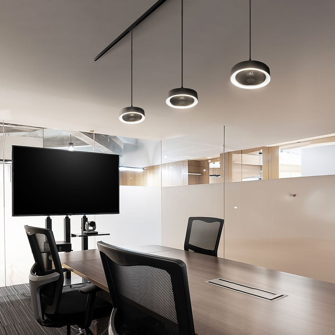 Czarna lampa LED z wiatrakiem i pilotem, zawieszona w biurze na szynie magnetycznej. Posiada okrągły kształt i emitującą światło obręcz. W tle widoczny telewizor na stojaku oraz biurko z krzesłami.