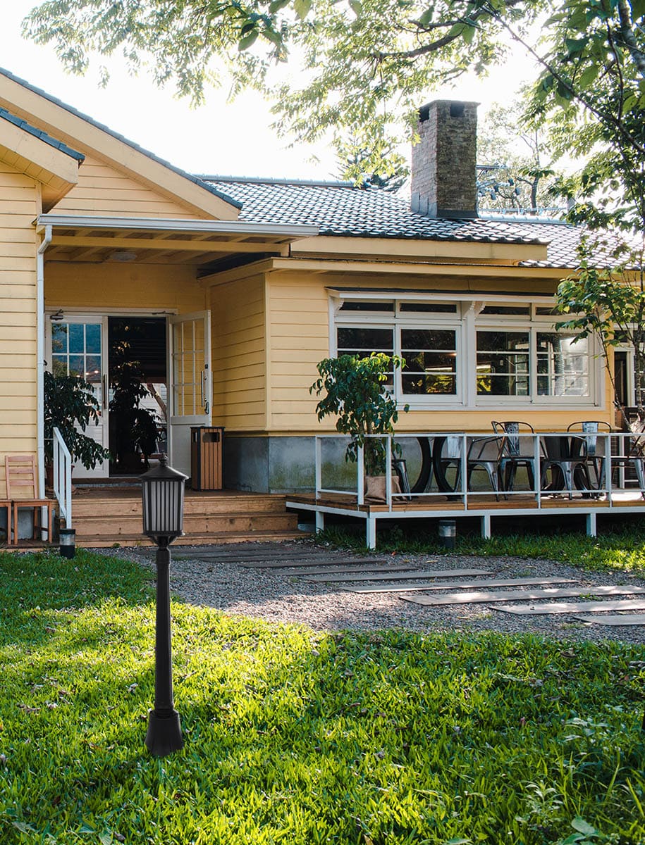 Lampa stojąca ogrodowa Detroit 1xE27 w kolorze czarnym, o prostym, cylindrycznym kształcie, umieszczona na trawniku przed żółtym domem. Widoczny detal klosza z poziomymi szczelinami oraz solidna podstawa. Tło przedstawia taras oraz roślinność.