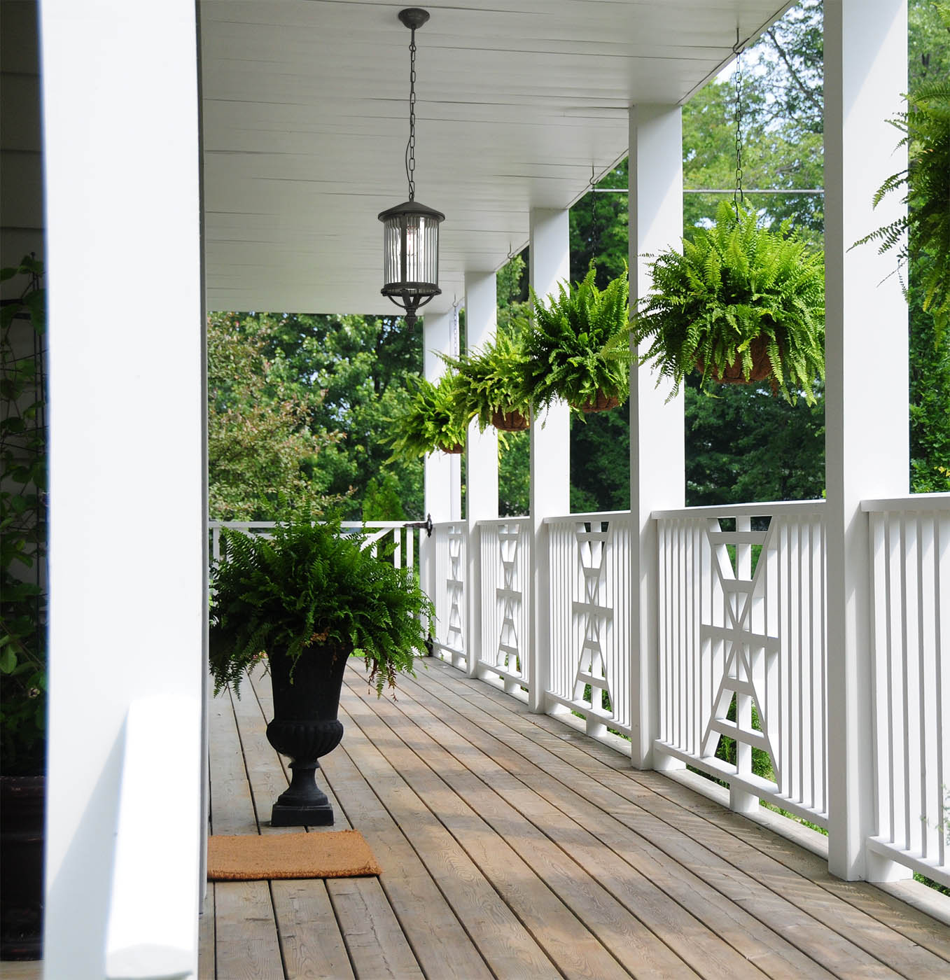 Lampa wisząca Miami w kolorze czarnym, z jednym gniazdem E27, zawieszona na łańcuchu. W tle drewniany taras z białymi balustradami i wiszącymi doniczkami z zielonymi paprociami. Jasne otoczenie z dużą ilością roślinności.