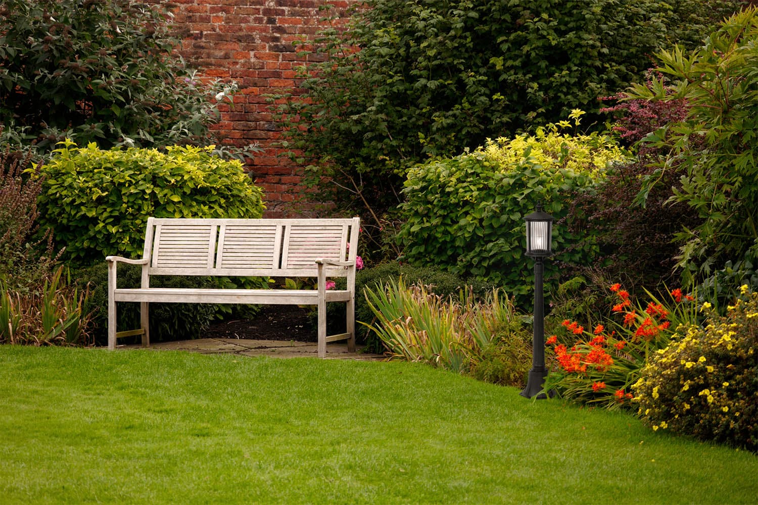 Lampa stojąca ogrodowa Miami, czarna, z miejscem na żarówkę E27. Ustawiona na trawniku obok drewnianej ławki, otoczona zielonymi krzewami i kolorowymi kwiatami. Wysoka, z dekoracyjnym kloszem, idealna do oświetlenia przestrzeni zewnętrznej.