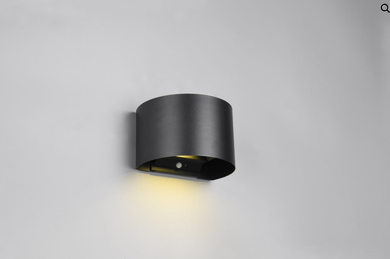 Kinkiet zewnętrzny w kształcie cylindrycznym, wykonany z czarnego metalu. Posiada czujnik ruchu i zmierzchu, emitujący światło LED o mocy 2x1W. Zamontowany na ścianie, skierowany w dół, z widocznym elementem czujnika. Tło jasnoszare.