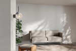 Kinkiet Vela 1xE14 w kolorze czarnym z bursztynowym kloszem, zamontowany na ścianie obok jasnej kanapy. Lampa ma zaokrąglony kształt klosza i stylowy, minimalistyczny design, otoczona roślinnością, co podkreśla nowoczesne wnętrze.